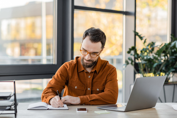 positive bearded businessman in eyeglasses and trendy shirt writing in notebook near laptop, smartphone with blank screen, documents tray and sticky notes at workplace in office - 写真・画像