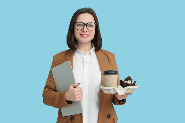 Happy young intern holding laptop, takeaway cup with hot drink and muffin on light blue background - Foto, imagen