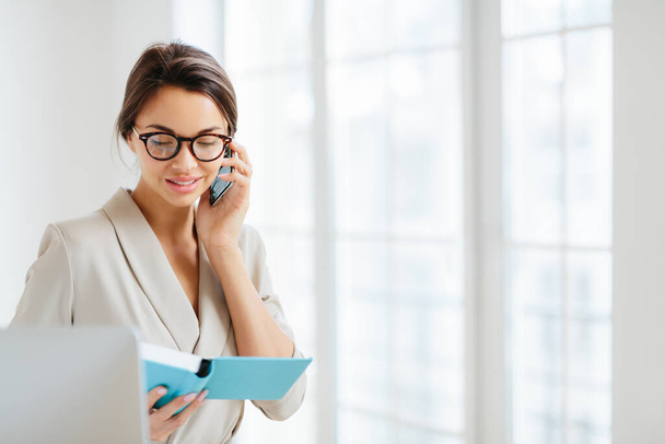 Horizontal view of business lady discusses working schedule with partner via modern smartphone, makes solution for project, focused in notepad, wears spectacles and formal suit, stands indoor - Foto, immagini