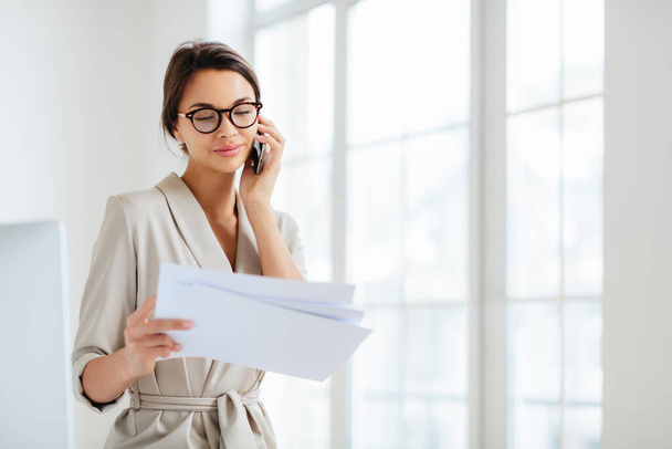 Horizontal shot of business lady discusses details of contract, works in office, concentrated on information from accountings, holds paper documents during telephone conversation, dressed formally - Zdjęcie, obraz