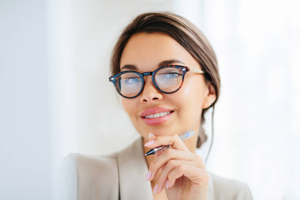 Pleasant looking young female model with makeup and manicure holds pen, touches chin gently, looks directly into camera, dressed in formal clothing, works in office of company, wears optical glasses - Foto, imagen