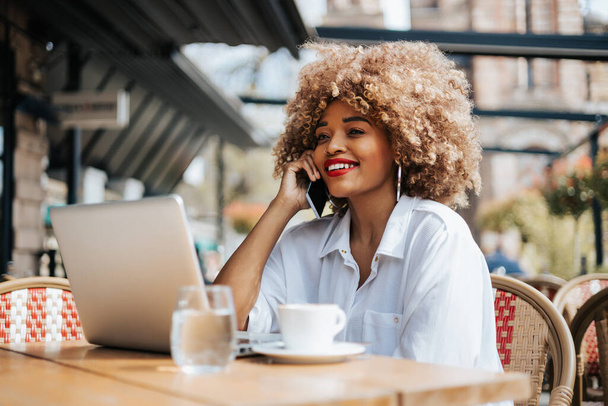 Beautiful black businesswoman sitting in restaurant or cafe bar and using her laptop computer for online communication or corporate video chat. Bright sunny day. - 写真・画像