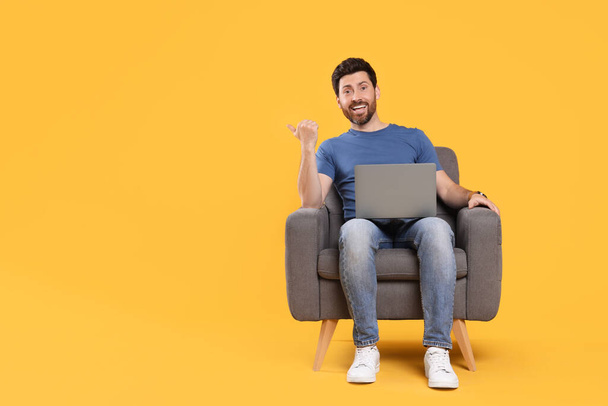 Emotional man with laptop sitting in armchair against yellow background. Space for text - Foto, Bild