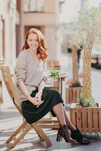 Photo of beautiful red haired woman dressed in stylish clothes, sits in open air, poses at cafeteria, has pleasant appearance, enjoys leisure time, smiles positively. People and lifestyle concept - Foto, immagini