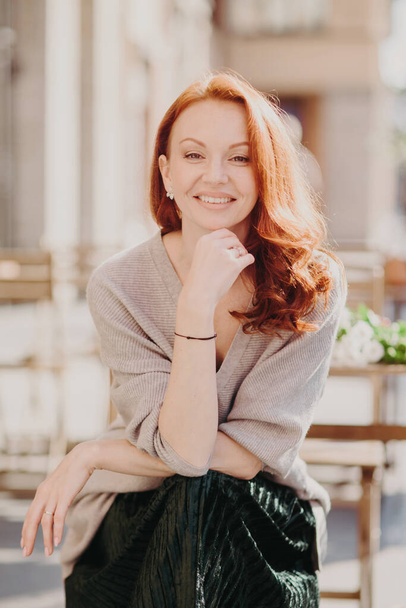Cheerful attractive red haired young woman has positive expression, keeps hand under chin, wears fashionable attire, looks direclty at camera, poses outside, being in high spirit, smiles happily - Photo, Image