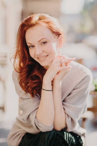 Image of lovely redhead female model smiles happily, has healthy skin, make up, keeps hands together, dressed in oversized jumper, poses outside, smiles gladfully. Having nice day or weekend - Фото, изображение