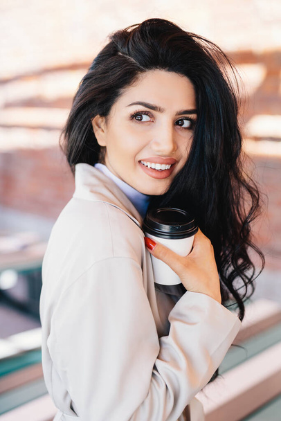Beautiful brunette lady with dark well-shaped eyebrows, appealing eyes and full lips smiling while looking aside showing her white perfect teeth drinking cappuccino in paper container posing outdoors - Foto, afbeelding