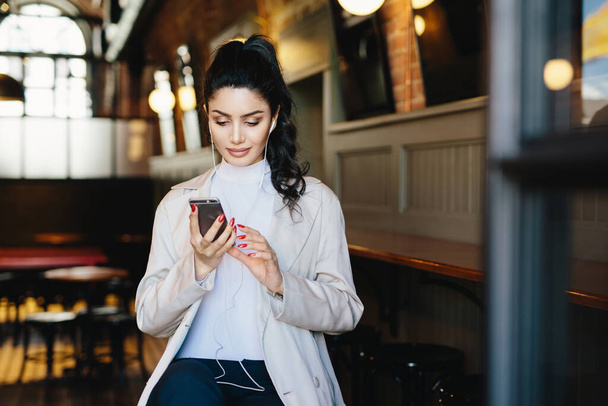 Pretty brunette woman with pony tail and appealing appearance sitting in cafe using her smartphone surfing internet using free wifi conncection and listening to her favourite music with earphones - Photo, Image