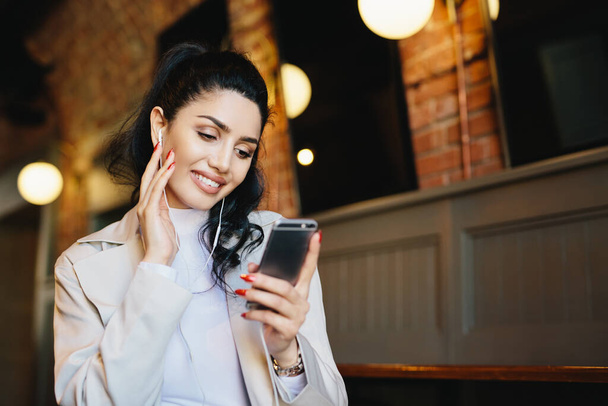 Smiling female with dark hair tied in pony tail wearing white clothes holding smartphone while listening to music on her electronic device. Stylish woman resting in cafe using app having smile on face - 写真・画像