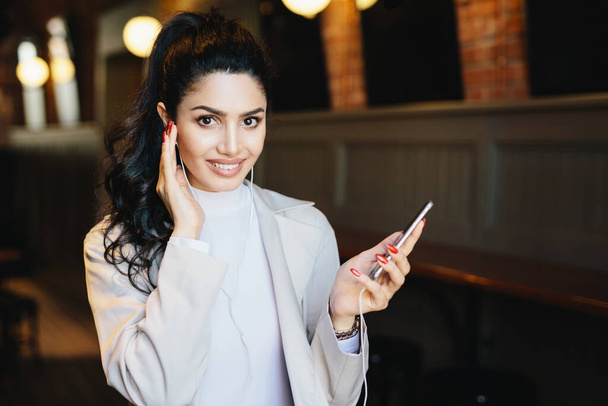 Cute woman with dark pony tail having long eyelashes and expressive eyes sitting at cafe listening to music online enjoying high-speed internet connection. People, beauty and technology concept - Photo, Image