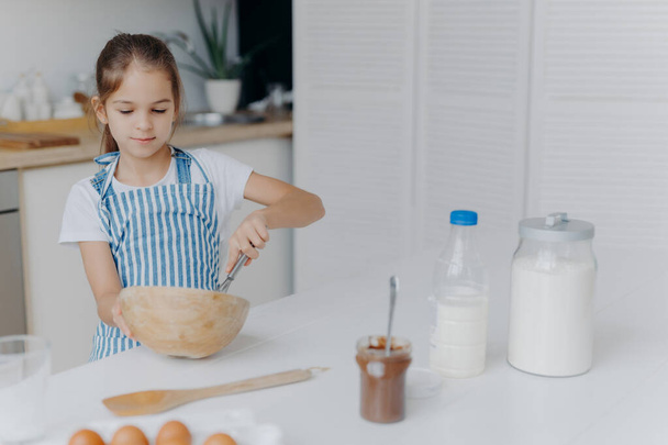 Children, cooking and home concept. Pretty dark haired girl whisks ingredients in bowl, busy preparing dough for cake, being future chef, wears white t shirt and striped apron, uses ingredients - Photo, Image