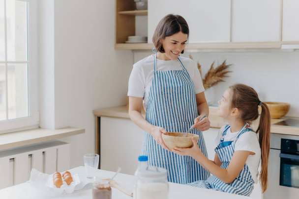 Indoor shot of caring mother with happy smile, cooks together with daughter in kitchen, prepare holiday pie, have pleasant talk, stand near table with different ingredients. Small helper helps mommy - Fotoğraf, Görsel