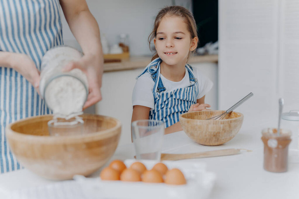 Curious little girl looks how mom prepares dough for pastry, learns to cook, gets culinary experience, wears apron. Faceless woman adds flour in bowl with other ingredients, pose in kitchen with child - Foto, imagen