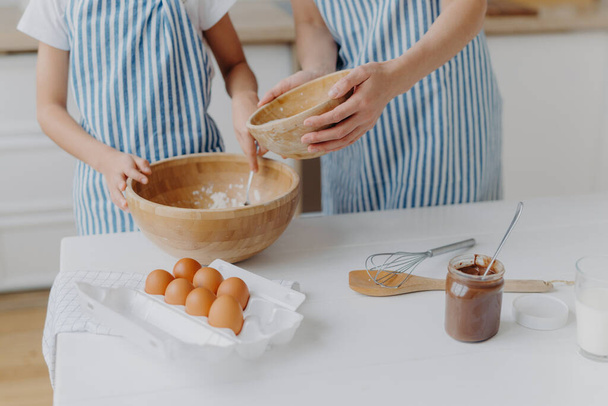 Cropped image of mothers and daughters hands mixing ingredients to prepare dough and bake tasty pastry, stand near kitchen table with eggs, melted chocolate in glass, wear striped blue aprons - Photo, Image