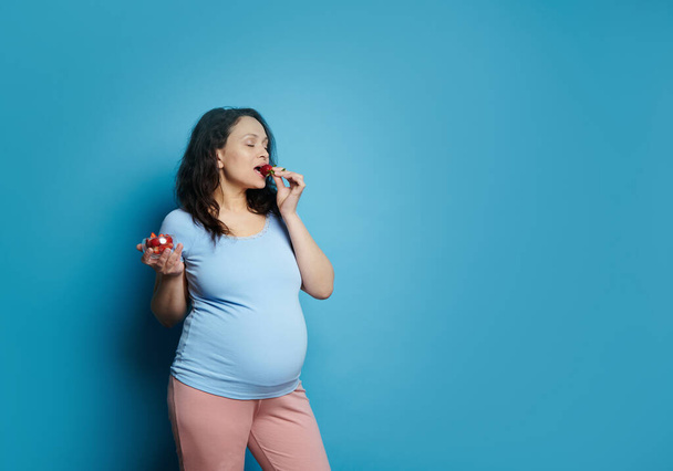 Delightful adult pregnant woman in blue t-shirt, eating delicious organic fresh sweet strawberries, isolated on blue background. Copy advertising space. Pregnancy. Healthy eating. Dieting concept - Photo, Image