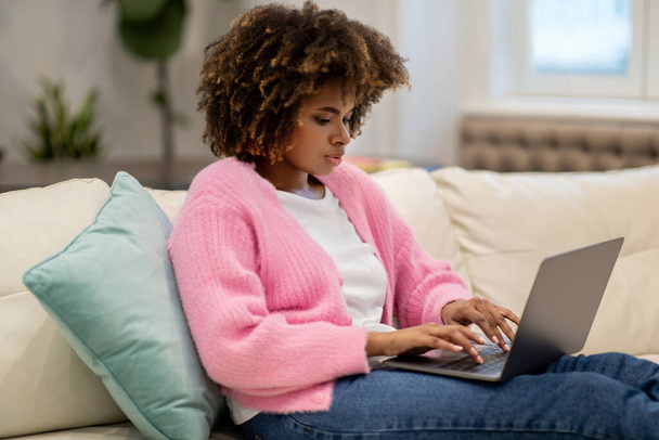 Nuori musta nainen yllään rento asu itsenäinen urakoitsija istuu sohvalla olohuoneessa, työskentelee kotoa, kannettavan tietokoneen avulla, kirjoittamalla tietokoneen näppäimistöllä, lähettämällä sähköposteja, kopioi tilaa - Valokuva, kuva