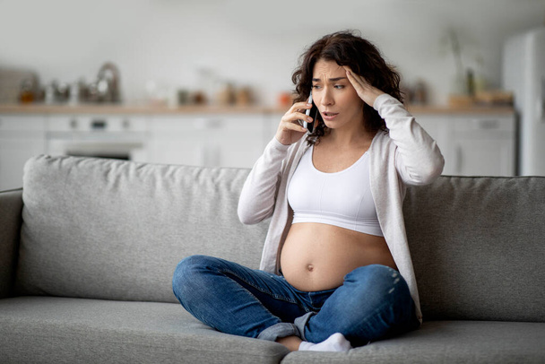 Mujer embarazada estresada hablando en el teléfono celular mientras está sentada en el sofá en casa, madre embarazada joven preocupada llamando al médico, tocando la cabeza y frunciendo el ceño, sintiéndose mal, espacio copiado - Foto, Imagen