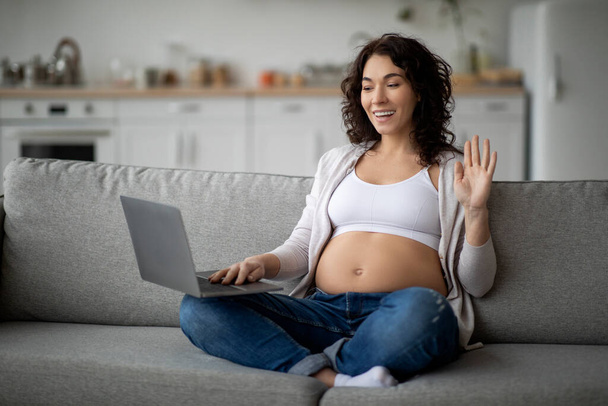 Communication en ligne. Femme enceinte souriante avec ordinateur portable faisant un appel vidéo à la maison tout en se reposant sur le canapé dans le salon, heureuse s'attendant mère agitant la main à la caméra Web, Copier l'espace - Photo, image