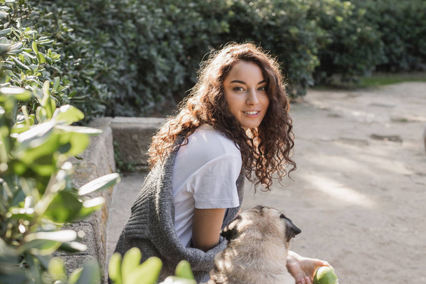 Carefree young curly woman in t-shirt and sweater holding green apple and looking at camera while sitting on stone bench near pug dog and relaxing in park in Barcelona, Spain  - Foto, afbeelding
