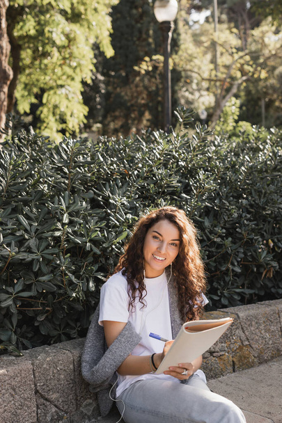 Femme souriante jeune et bouclée en vêtements décontractés utilisant des écouteurs filaires, écrivant sur un carnet et regardant la caméra alors qu'elle était assise sur un banc en pierre dans un parc à Barcelone, Espagne  - Photo, image