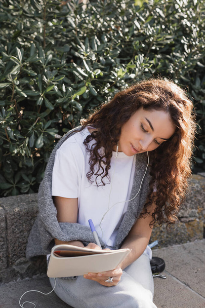 Молодая и кудрявая женщина в повседневной одежде слушает музыку в проводных наушниках и пишет на ноутбуке, расслабляясь на каменной скамейке в парке в Барселоне, Испания  - Фото, изображение