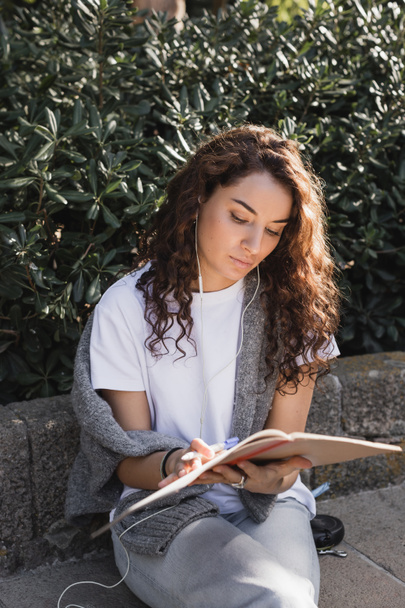 Young curly and pretty woman in casual clothes and wired earphones looking at notebook and holding marker while sitting on stone bench in park in Barcelona, Spain  - Photo, Image