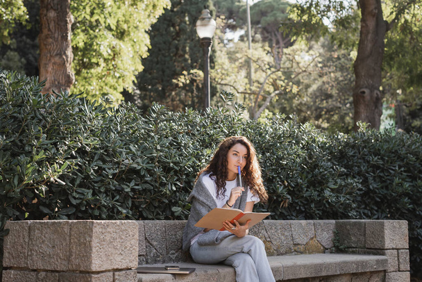 Töprengő fiatal és göndör nő alkalmi ruhák kezében notebook és marker ajkak közelében, miközben ül eszközök mellett kő pad és zöld növények parkolóban nappal Barcelonában, Spanyolország  - Fotó, kép