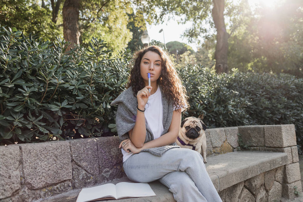 Mulher jovem e encaracolada focada em roupas casuais segurando marcador e olhando para a câmera perto do notebook com espaço de cópia e cachorro sentado no banco de pedra no parque em Barcelona, Espanha  - Foto, Imagem
