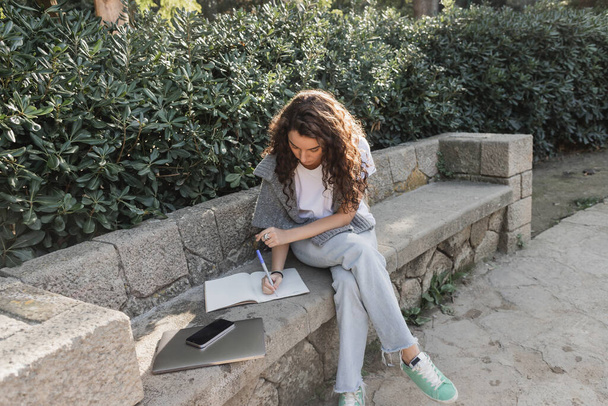 Young and curly woman in warm sweater on shoulders writing on notebook near laptop and smartphone with blank screen on stone bench beside green bushes in park in Barcelona, Spain  - Photo, Image