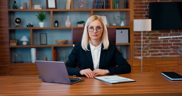 Retrato de encantadora mujer de negocios rubia adulta concentrada en ropa formal que se sienta delante de la cámara en la oficina moderna - Foto, Imagen