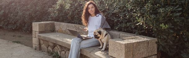 Mladá a kudrnatá hezká žena pomocí gadgets a mazlení pug dog při pohledu na kameru a trávení času na kamenné lavičce v zeleném parku v Barceloně, Španělsko, prapor  - Fotografie, Obrázek