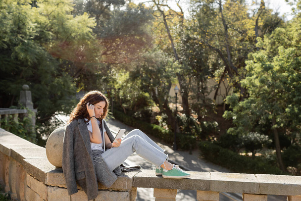Joven y rizado freelancer con chaqueta abrigada usando smartphone y auriculares mientras sostiene el portátil y se sienta en la valla de piedra en el borroso parque de verano en Barcelona, España  - Foto, imagen