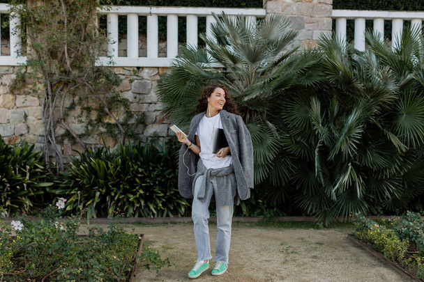 Positive junge und lockige Frau mit Kopfhörern und warmer Jacke, die Gadgets in der Hand hält und wegschaut, während sie tagsüber im Park in der Nähe von Palmen spaziert  - Foto, Bild