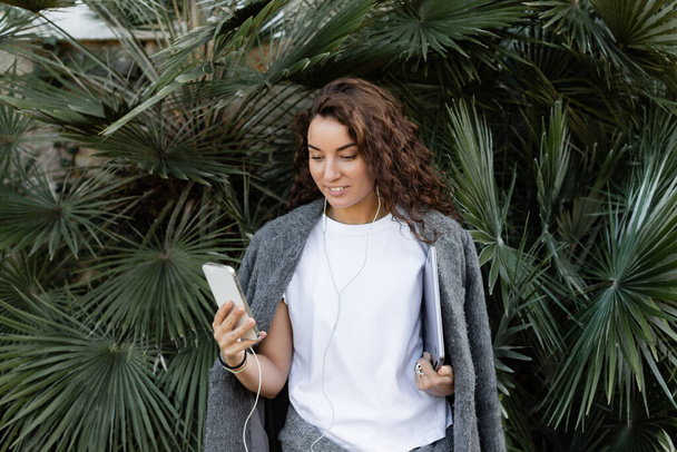 Cheerful young and curly freelancer in warm jacket using earphones and smartphone while holding laptop and standing near green palm trees in park in Barcelona, Spain  - Photo, Image
