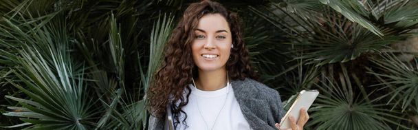 Positive and young curly freelancer in earphones and casual clothes holding gadgets and looking at camera while standing near palm trees in park at daytime in Barcelona, Spain, banner  - Photo, Image