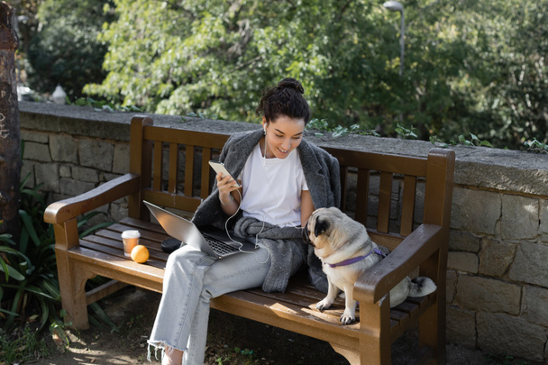 Freelancer sonriente en audífonos sosteniendo smartphone y mirando cachorro mientras está sentado cerca del portátil, café para llevar y naranja en un banco de madera y pasando tiempo en el parque en Barcelona, España  - Foto, Imagen