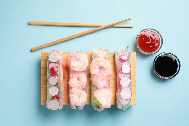 Different delicious spring rolls, chopsticks and sauces on light blue background, flat lay - Foto, Imagem