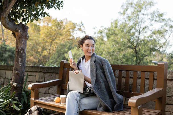 Uśmiechnięta brunetka freelancer w ciepłej kurtce przy użyciu słuchawek i urządzeń podczas spędzania czasu w pobliżu mopsa, kawa na wynos i pomarańczowy na drewnianej ławce w parku w Barcelonie, Hiszpania  - Zdjęcie, obraz
