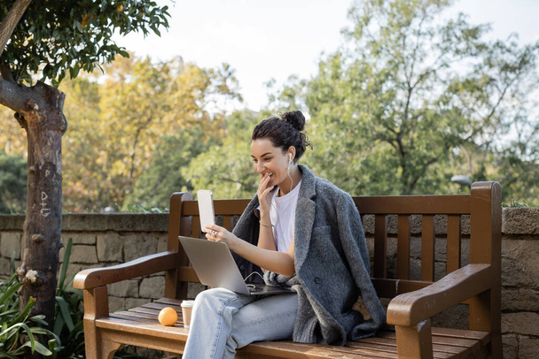 Smiling young freelancer in warm jacket and earphones smiling, covering mouth, having video call on smartphone near laptop and coffee to go on wooden bench in park in Barcelona, Spain  - Photo, Image