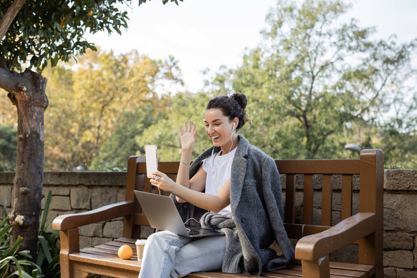 Cheerful young freelancer in warm jacket and earphones waving hand at smartphone while having video call on smartphone near laptop, coffee to go and orange on wooden bench in park in Barcelona, Spain  - Fotoğraf, Görsel