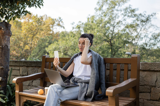 Kudrnatý a pěkný na volné noze v teplé bundě s videohovor na smartphone v blízkosti notebooku, káva jít a pomeranč na dřevěné lavičce v parku ve dne v Barceloně, Španělsko  - Fotografie, Obrázek