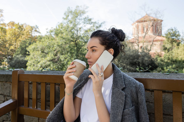 Güzel ve kıvırcık tişörtlü ve kalın ceketli bir kadın akıllı telefondan konuşuyor ve elinde kahve tutarken Barselona, İspanya 'da parktaki ahşap bankta oturuyor, sıcak içecekle kağıt bardak içiyor.  - Fotoğraf, Görsel