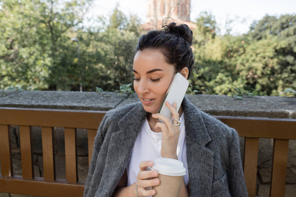 Обережна молода жінка в теплому куртці говорить по мобільному телефону і тримає каву, щоб піти, проводячи час на вулиці на дерев'яній лавці в парку вдень в Барселоні (Іспанія).  - Фото, зображення