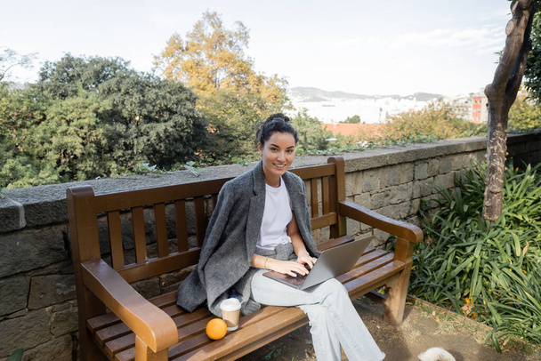 Joven freelancer sonriente con chaqueta abrigada y ropa casual mirando a la cámara, usando un portátil y sentado cerca del café para ir y naranja en un banco de madera en el parque en Barcelona, España  - Foto, Imagen