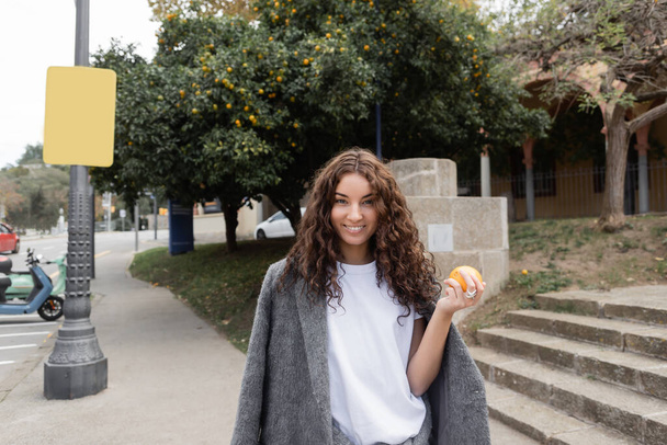 Uśmiechnięta młoda i kręcona kobieta w ciepłej kurtce trzymająca świeżą pomarańczę i patrząca w kamerę stojąc na rozmytej miejskiej ulicy w tle w Barcelonie, Hiszpania, lampa uliczna, skuter motorowy - Zdjęcie, obraz