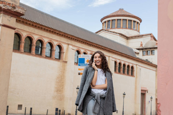 Smiling young and curly woman in warm jacket and casual clothes talking on smartphone and looking away while standing near historic landmark on urban street in Barcelona, Spain, ancient building - Fotografie, Obrázek