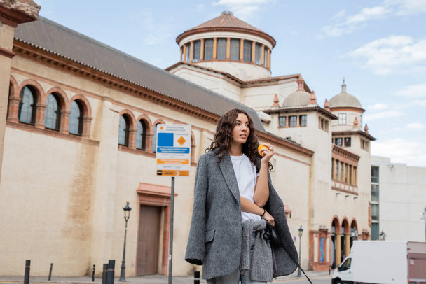 Young curly woman in grey casual jacket holding fresh orange and leash while looking away with blurred historical landmark at background in Barcelona, Spain, ancient building  - Φωτογραφία, εικόνα