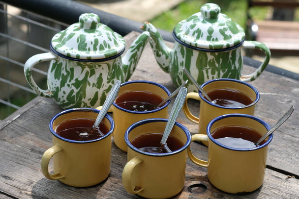 Teh Poci Gula Batu, Teko Blirik. jest gorąca herbata słodzona cukrem kamiennym, serwowane z zabytkowym czajnikiem emalii i kubek przeciwnika na brązowym drewnianym rustykalnym stole. Napoje indonezyjskie. - Zdjęcie, obraz