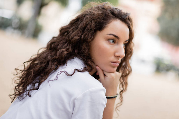 Retrato de una joven mujer rizada y pensativa en camiseta blanca mirando hacia otro lado y cogida de la mano cerca de la barbilla mientras está parada en la borrosa calle urbana de Barcelona, España  - Foto, imagen