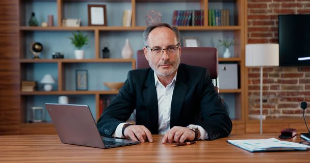 Portrait of attractive confident experienced purposeful bearded ceo in business suit which posing on camera during his work on laptop in modern office - 写真・画像